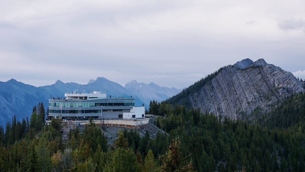 Banff Gondola summit building on Sulphur Mountain above Banff, Alberta, Canada, surrounded by evergreen forest with steep rocky ridges and layered Canadian Rockies extending into the distance.