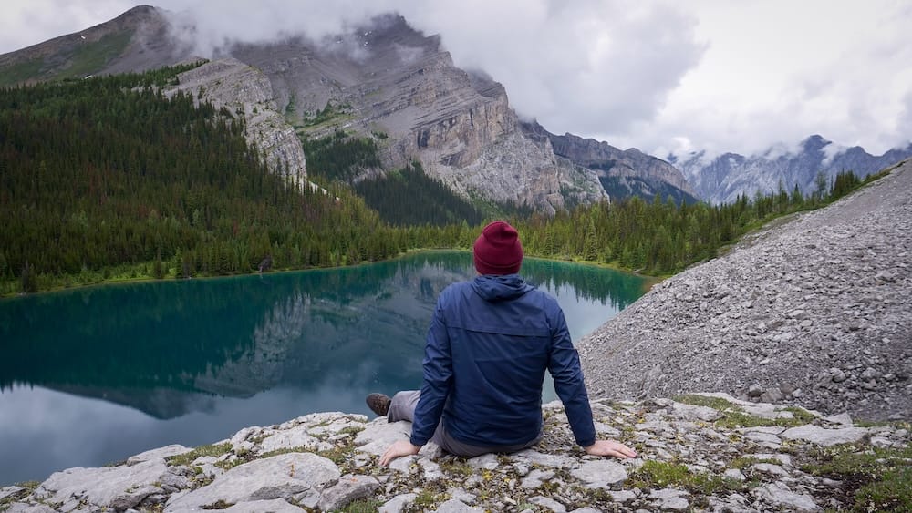Samuel Jeffery overlooking an alpine lake in Banff National Park, Alberta, Canada, beneath cloudy mountain peaks Samuel Jeffery sits on a rocky ledge above a turquoise alpine lake in Banff National Park, Alberta, Canada, with forested slopes, rugged Rocky Mountain peaks and low clouds behind him.