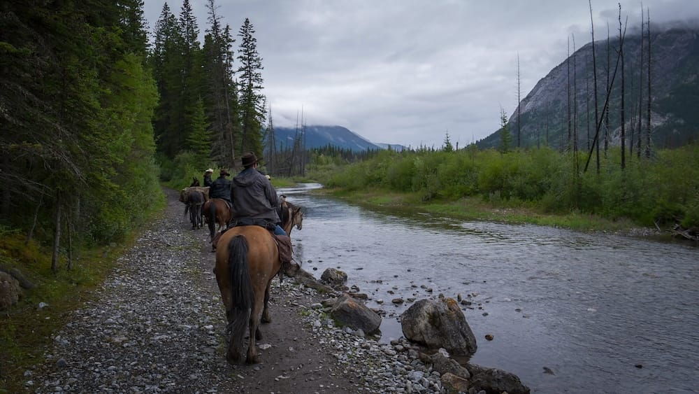 A group of horseback riders follows a narrow riverside trail near Banff town in Alberta, Canada, with forest, rocky shoreline and steep mountain slopes beneath a cloudy sky.