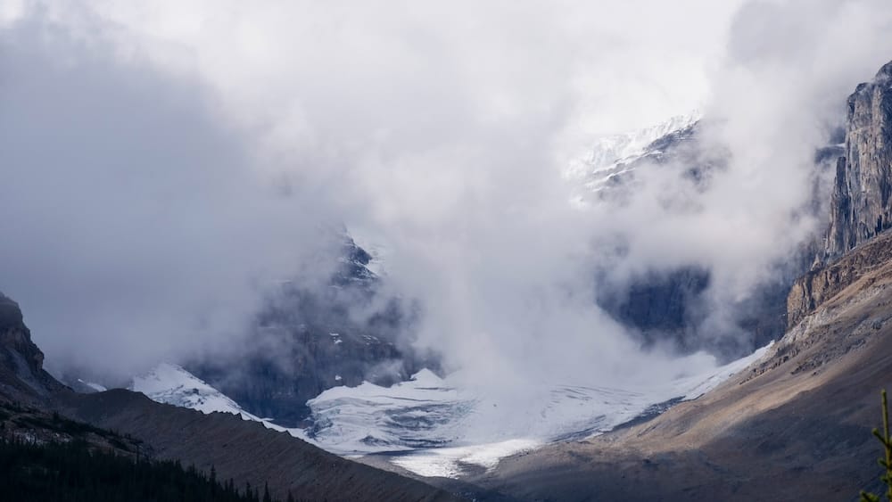 Moody mountain weather in Banff National Park, Alberta, Canada, with clouds, snow and glaciated Rocky Mountain peaks Low clouds and mist wrap around snow-covered slopes and a glacier in Banff National Park, Alberta, Canada, creating a dramatic Rocky Mountain scene with steep rock walls and shifting mountain weather.