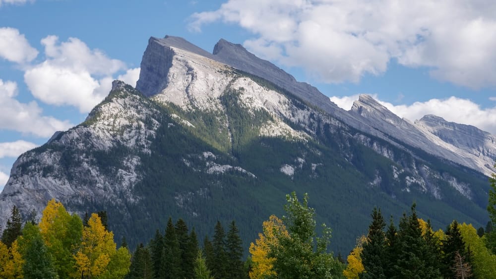 Steep gray Rocky Mountain peaks rising above dense evergreen forest and yellow autumn foliage in Banff National Park, Alberta, Canada, under a blue sky with scattered clouds.
