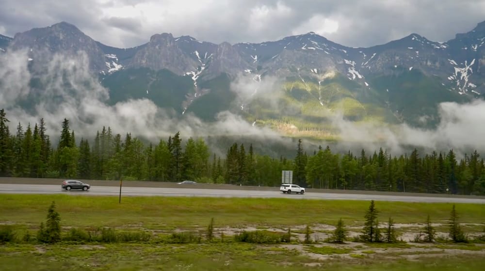 Scenic drive through Banff National Park, Alberta, Canada with mountain highway, forest and low cloud cover Vehicles travel along a highway in Banff National Park, Alberta, Canada, beneath forested mountain slopes with low clouds and mist drifting across the Canadian Rockies peaks.