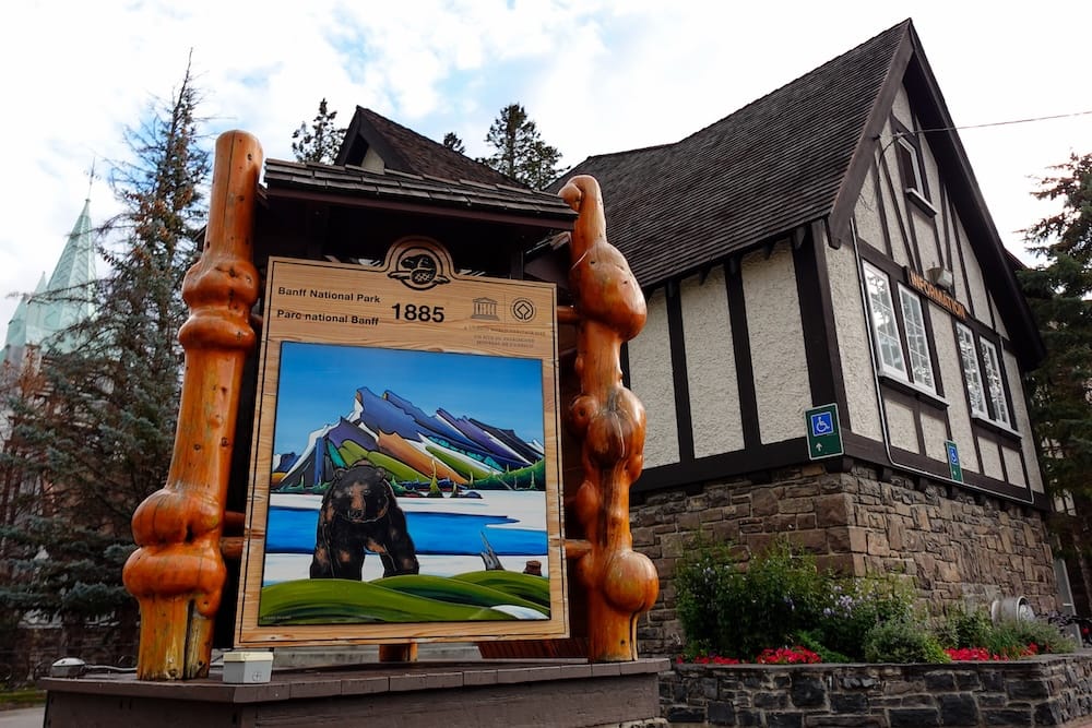 Wooden Banff National Park sign in Banff town, Alberta, Canada, displaying 1885 and colourful bear-and-mountain artwork beside a historic-style building.