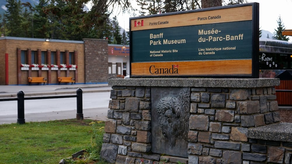 Parks Canada sign for Banff Park Museum National Historic Site in Banff, Alberta, Canada, mounted on a stone base beside the street with trees and town buildings behind it.