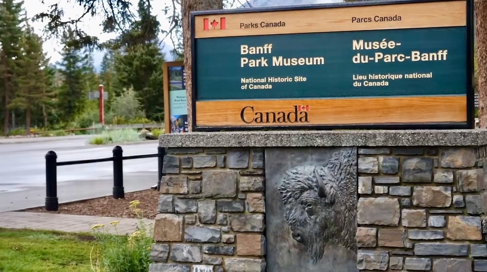 Parks Canada sign for Banff Park Museum in Banff town, Alberta, Canada, mounted on a stone base with a bison relief and trees and mountain scenery visible behind it.