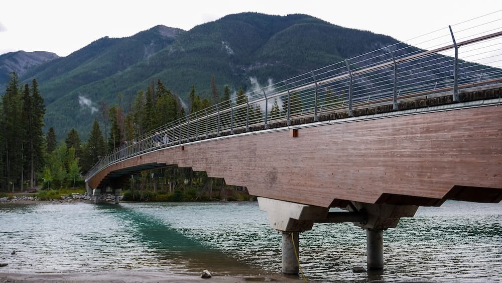 A pedestrian bridge crossing the Bow River in Banff town, Alberta, Canada, with turquoise water, evergreen forest and steep Canadian Rockies mountains rising behind it.