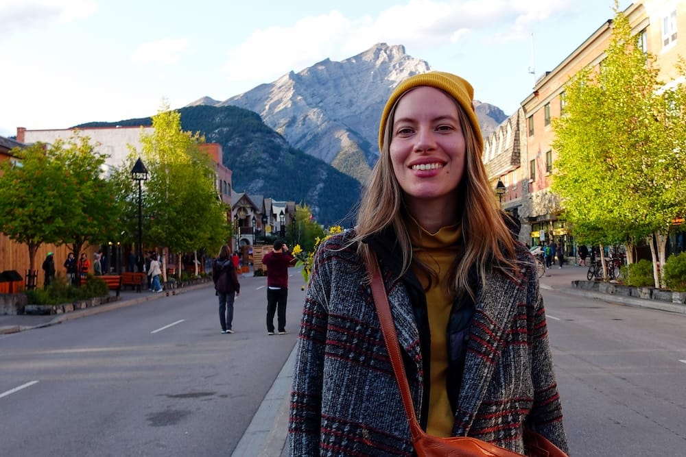 Audrey Bergner in Banff Town, Alberta, Canada, with Downtown Streets and Canadian Rockies Mountain Views Audrey Bergner smiling on a downtown street in Banff town, Alberta, Canada, with shops, leafy trees, pedestrians and a steep Canadian Rockies mountain rising directly behind her.