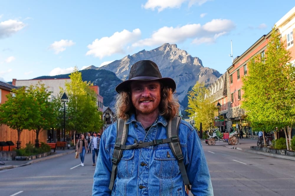 Samuel Jeffery in downtown Banff, Alberta, Canada, with shops, trees and Rocky Mountain scenery behind him Samuel Jeffery stands on a street in Banff town, Alberta, Canada, wearing a backpack and hat, with shops, leafy trees and a dramatic Rocky Mountain peak rising behind the downtown streetscape.