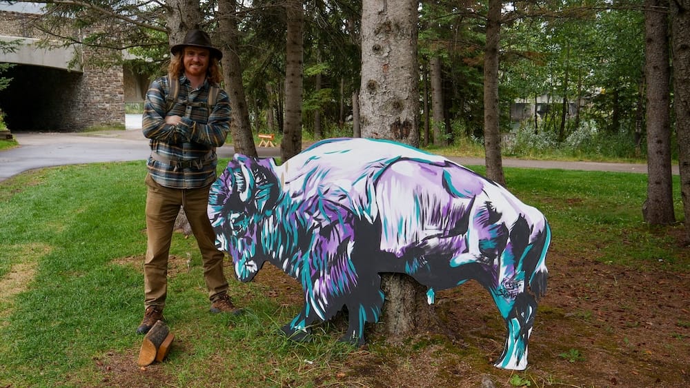 Samuel Jeffery standing beside a colourful painted bison sculpture in Banff, Alberta, Canada, with evergreen trees, grass and a quiet pedestrian area surrounding the public artwork.