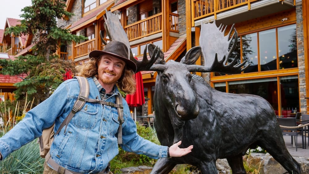 Samuel Jeffery posing beside a large moose statue in Banff town, Alberta, Canada, with timber-and-stone buildings and outdoor seating in the background.