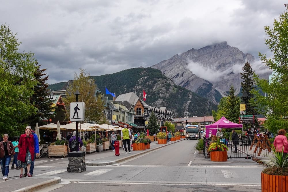 Banff town street scene in Alberta, Canada with pedestrians, shops, transit and Rocky Mountain backdrop A street scene in Banff town, Alberta, Canada, with pedestrians, shops, outdoor seating, transit and colorful planters beneath a large grey Rocky Mountain partly wrapped in low cloud.
