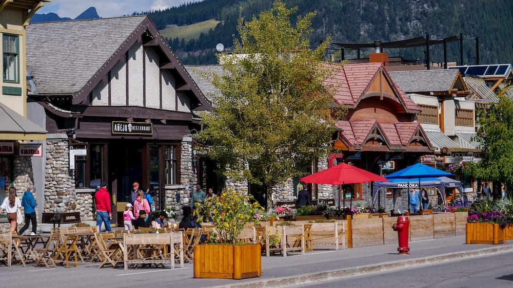 Downtown Banff Town Street Scene in Alberta, Canada, with Restaurant Patios and Canadian Rockies Backdrop Street scene in Banff town, Alberta, Canada, with restaurant patios, colourful umbrellas, pedestrians, chalet-style buildings and forested Canadian Rockies slopes rising behind downtown.