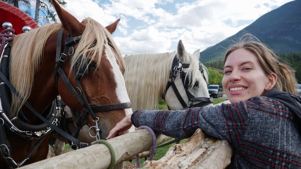 Caballos de Banff Trail Riders antes del paseo en carreta en Banff, Alberta, Canadá, con Audrey Bergner Audrey Bergner se apoya en una cerca de madera junto a dos caballos con arnés antes del paseo en carreta de Banff Trail Riders en Banff, Alberta, Canadá, con montañas y cielo parcialmente nublado de fondo.