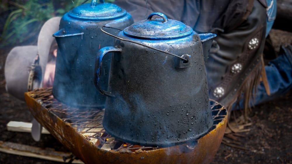 Café al fuego durante una cabalgata de Banff Trail Riders en el Parque Nacional Banff, Alberta, Canadá Dos cafeteras de esmalte azul están sobre una parrilla al fuego durante una cabalgata de Banff Trail Riders en el Parque Nacional Banff, Alberta, Canadá, con humo, botas y equipo de campamento alrededor.