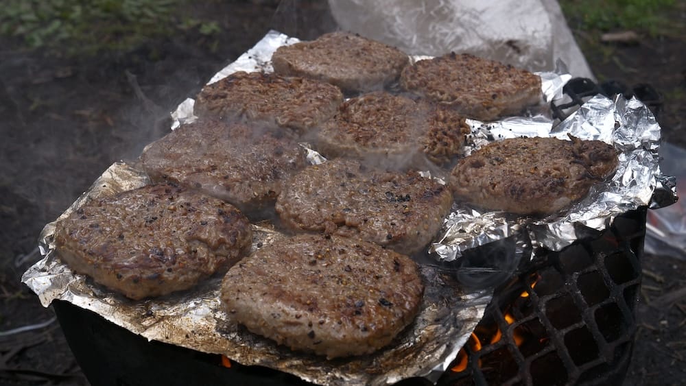 Hamburguesas a la parrilla durante un almuerzo de Banff Trail Riders en el Parque Nacional Banff Varias hamburguesas se cocinan sobre una parrilla al fuego durante una cabalgata de Banff Trail Riders en el Parque Nacional Banff, Alberta, Canadá, con humo elevándose sobre la superficie cubierta con papel aluminio.