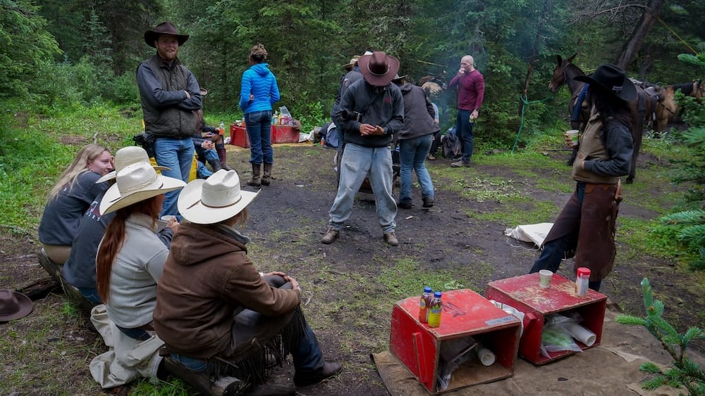 Backcountry lunch stop with Banff Trail Riders guests and horses in Banff National Park, Alberta, Canada Banff Trail Riders guests gather for a backcountry lunch stop in Banff National Park, Alberta, Canada, with riders seated in a forest clearing, food supplies nearby and saddled horses tied among the trees.
