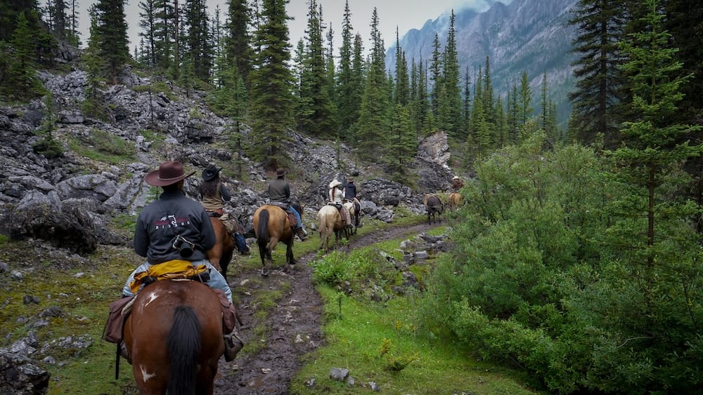 Horseback riding through forest and mountain terrain in Banff National Park, Alberta, Canada with Banff Trail Riders A small group of horseback riders follows a muddy backcountry trail through evergreen forest in Banff National Park, Alberta, Canada, with rocky slopes and steep mountain walls visible ahead.