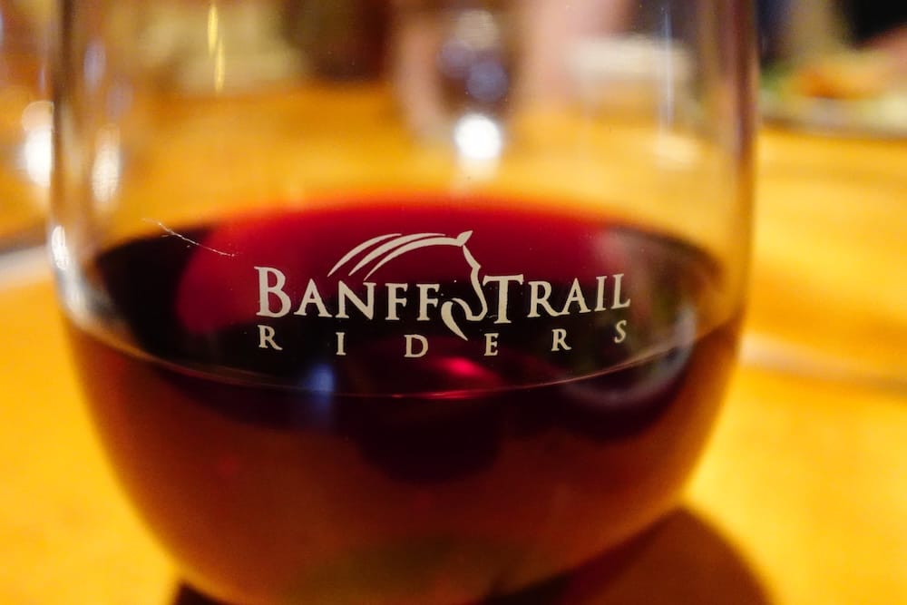 Vino tinto de Banff Trail Riders durante una cena en un lodge del Parque Nacional Banff, Alberta, Canadá Una copa de vino tinto con el logo de Banff Trail Riders está sobre una mesa de madera durante una travesía a caballo en el Parque Nacional Banff, Alberta, Canadá, con el ambiente de cena del lodge desenfocado detrás.