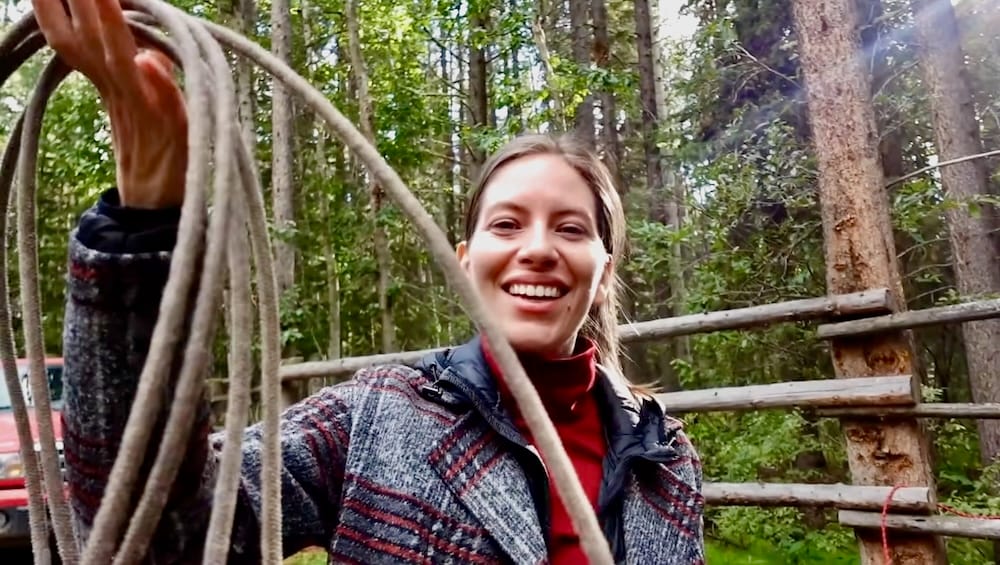 Audrey Bergner probando el lazo durante el Banff Wagon Ride With Cowboy Cookout en Alberta, Canadá Audrey Bergner sonríe mientras sostiene un lazo durante el paseo en carreta con Cowboy Cookout en el Parque Nacional Banff, Alberta, Canadá, con cercas de madera y bosque detrás.