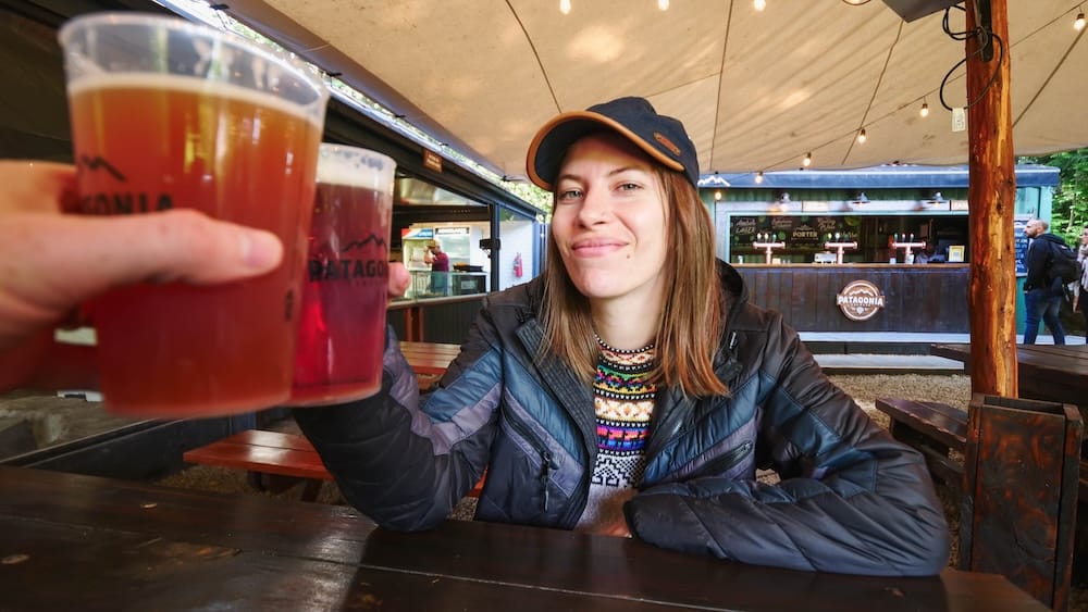 Audrey Bergner and Samuel Jeffery toasting plastic cups of Cervecería Patagonia craft beer in Bariloche, Argentina, capturing the shared reward of arriving at this Lake District brewery after navigating transport, timing, and the journey out of town.
