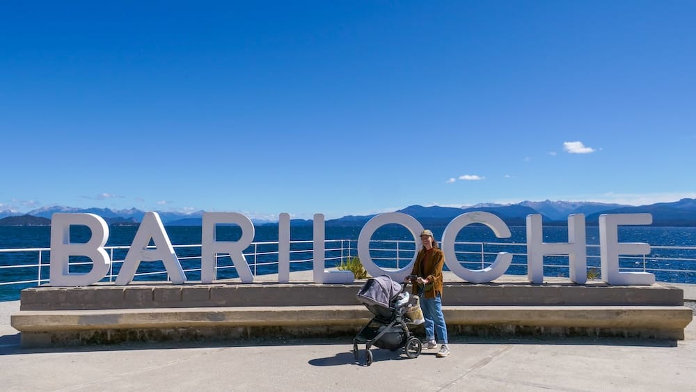 Audrey Bergner and Aurelia Jeffery at the Bariloche sign in Río Negro, Patagonia, Argentina Audrey Bergner stands with Aurelia Jeffery in a stroller beside the large Bariloche landmark sign, with Nahuel Huapi Lake and distant mountains behind them in Río Negro, Patagonia, Argentina.
