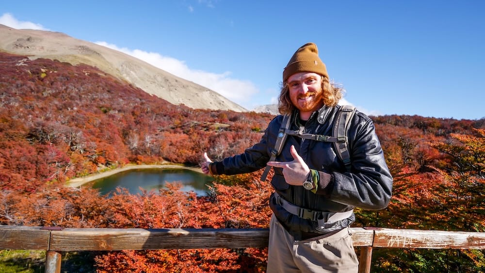 Bariloche Mirador Laguna Verde in autumn with Samuel Jeffery pointing toward a small lake, red lenga forest, mountain slopes, and seasonal hiking scenery during Project 23 fieldwork in Argentine Patagonia.
