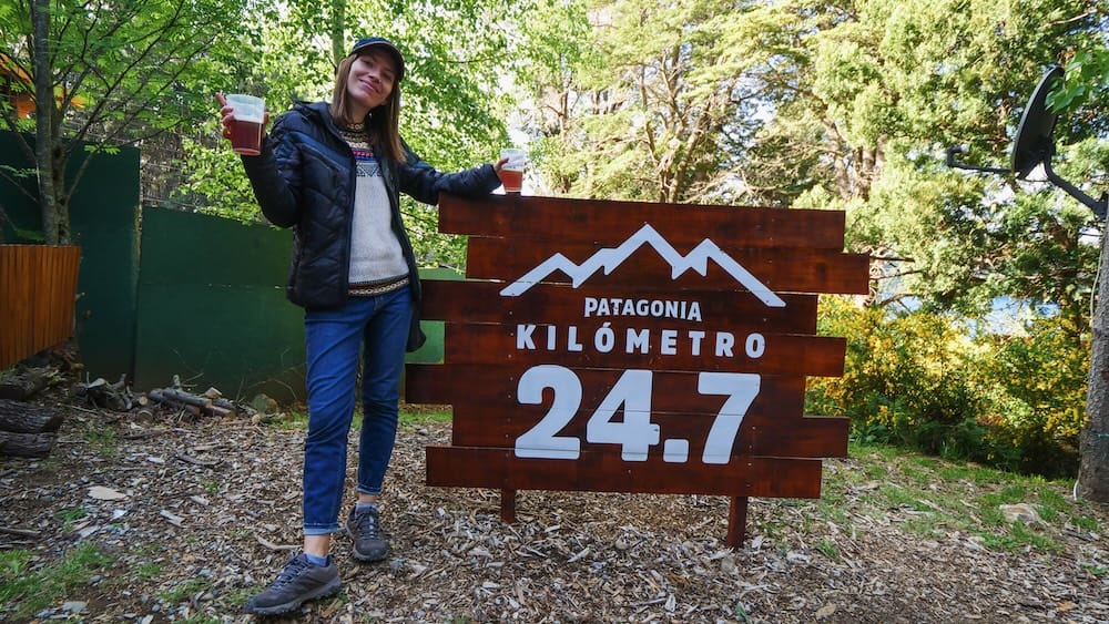 Audrey Bergner holding two plastic cups of craft beer beside the Patagonia Kilómetro 24.7 sign in Bariloche, Argentina, highlighting the destination brewery experience where travelers venture outside town for lake views, fresh pours, and a well-earned post-journey reward.
