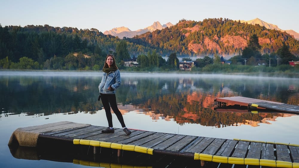 Bariloche Patagonia morning lake reflections with Audrey Bergner on dock Audrey Bergner standing on a wooden dock at a calm lake in Bariloche Patagonia Argentina with morning mist, forested hills, and Andes mountains reflected in the water