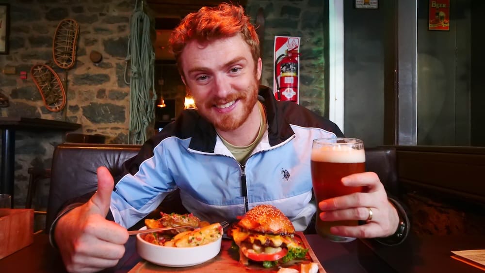 Samuel Jeffery holding a pint of craft beer and a gourmet burger at Cervecería Manush in Bariloche, Patagonia, Argentina, capturing the classic post-hike reward culture where hearty food and local beer go hand in hand in the Lake District.
