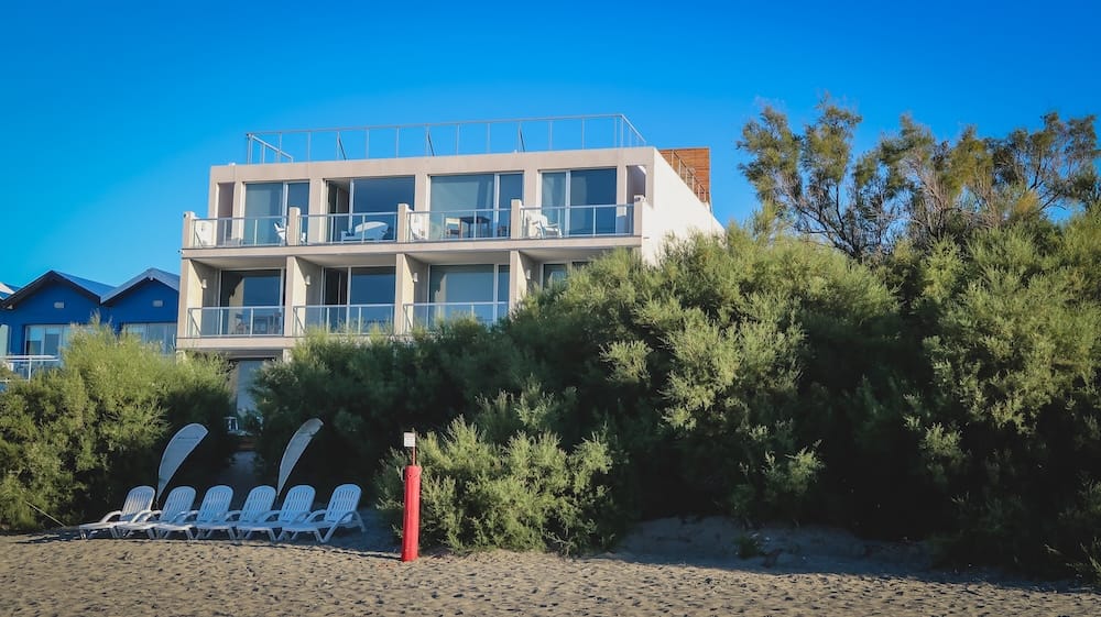 Un alojamiento moderno frente a la playa se eleva detrás del matorral costero y la arena en Puerto Pirámides, Península Valdés, Chubut, Patagonia, Argentina, con balcones, reposeras y cielo azul.