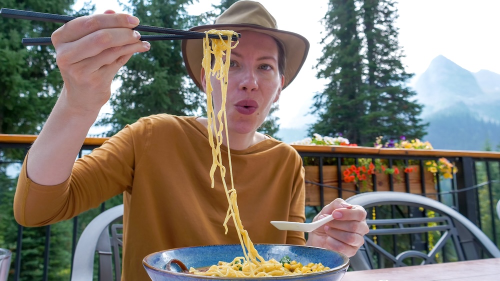 Bear Lodge Bistro patio at Island Lake Lodge in Fernie, British Columbia, as Audrey Bergner of That Backpacker lifts miso ramen noodles with chopsticks during a relaxed lunch stop, with alpine forest and mountain views framing the outdoor dining experience.
