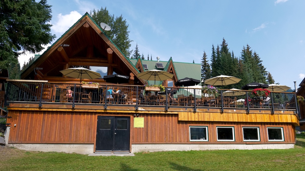 Bear Lodge Bistro outdoor patio at Island Lake Lodge in Fernie, British Columbia, featuring umbrella-covered tables, log lodge architecture, and forest surroundings where guests enjoy lunch and drinks with mountain air and relaxed alpine lodge vibes.
