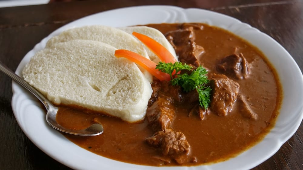 Un plato hondo de goulash de carne con trozos tiernos en una salsa marrón espesa, servido con dos rodajas de dumpling de pan, perejil fresco y tiras de morrón en Bistro Praha, un restaurante checo en el centro de Edmonton, Alberta.
