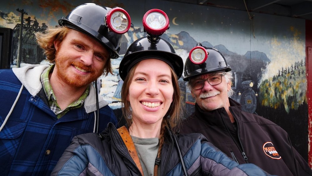 Bellevue Underground Mine Tour in Crowsnest Pass, Alberta, with Nomadic Samuel Jeffery and Audrey Bergner and Daniel Bergner wearing hard hats and headlamps beside a mining mural—an easy Fernie, BC day trip for history lovers.
