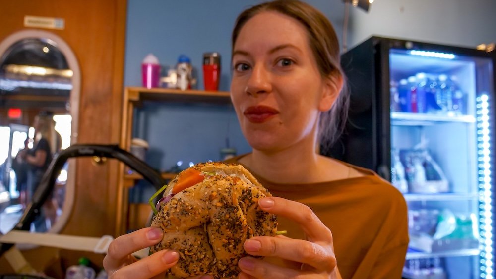 Audrey enjoying the Smoked Salmon Stack at Big Bang Bagels Audrey Bergner of That Backpacker holds a Smoked Salmon Stack bagel at Big Bang Bagels in Fernie, British Columbia, featuring cream cheese, alfalfa sprouts, red onion, and classic West Coast flavours inside a fresh everything bagel as she enjoys breakfast in the cozy café.