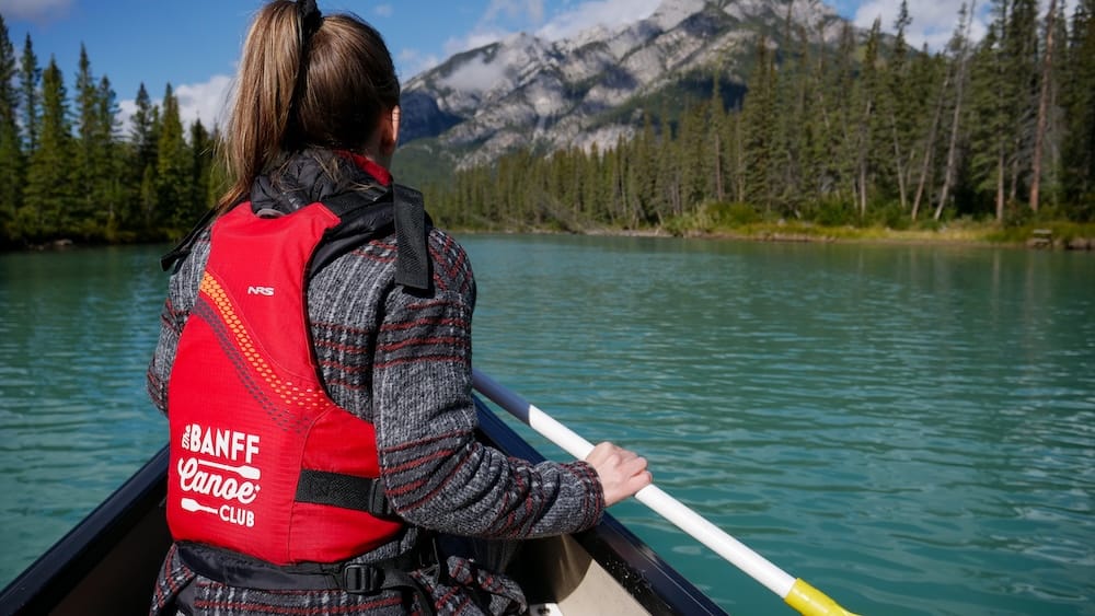 Tour en canoa grande, río Bow, Banff, Alberta, Canadá – Audrey Bergner Audrey Bergner remando desde la proa de una canoa del Banff Canoe Club en el río Bow cerca de Banff, Alberta, Canadá, con una montaña y orillas bordeadas de pinos por delante.