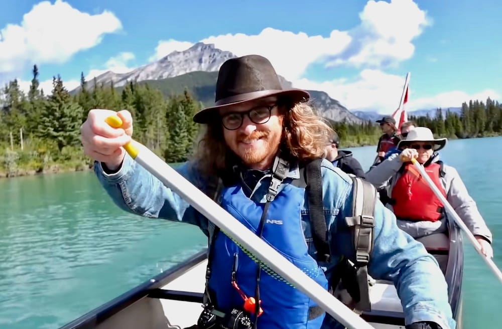 Samuel Jeffery remando contra la corriente junto a otros pasajeros a bordo del Big Canoe Tour compartido en el río Bow cerca de Banff, Alberta, Canadá, con una montaña y un bosque de pinos detrás de él.