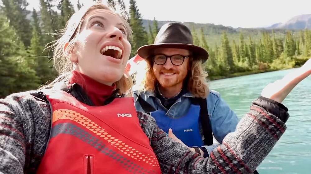 Audrey Bergner y Samuel Jeffery sonriendo y riendo con chalecos salvavidas durante el Big Canoe Tour en el río Bow cerca de Banff, Alberta, Canadá, con un bosque de pinos en la orilla a sus espaldas.