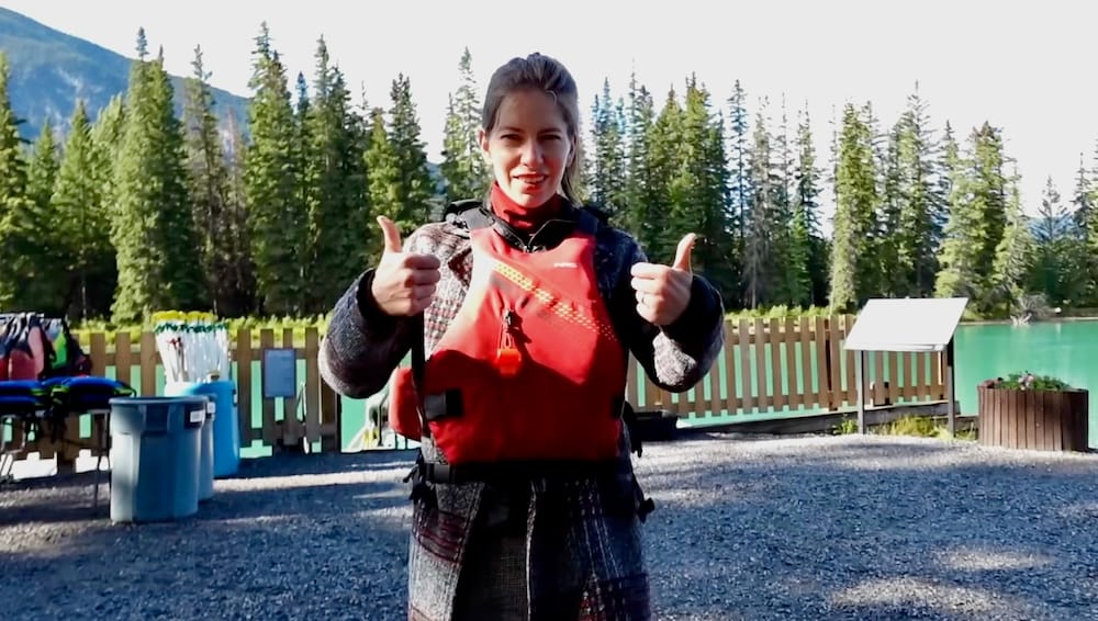 Audrey Bergner con el pulgar hacia arriba y chaleco salvavidas en el muelle del Banff Canoe Club en el río Bow en Banff, Alberta, Canadá, con tachos para el equipo y un cartel informativo visibles detrás de ella.