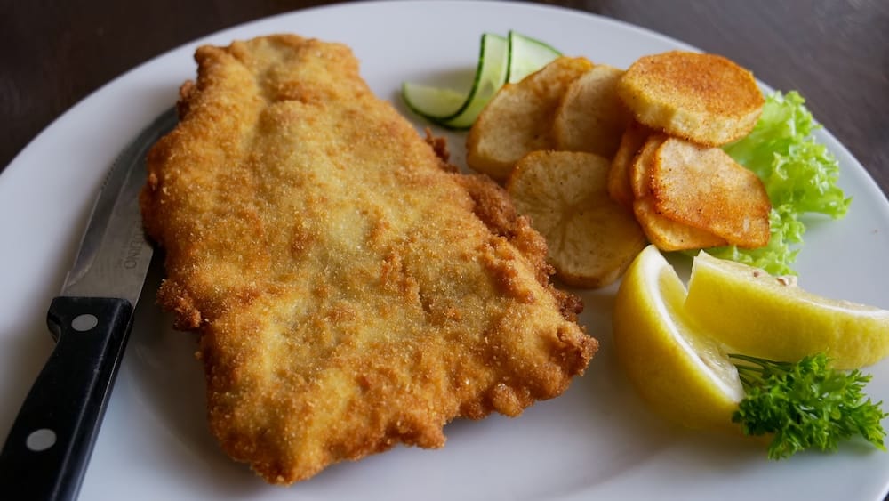 Schnitzel with Fried Potatoes at Bistro Praha in Edmonton, Alberta, Canada Breaded schnitzel at Bistro Praha in Edmonton, Alberta, Canada, served with fried potato slices, lemon wedges, cucumber, lettuce and parsley on a white plate.