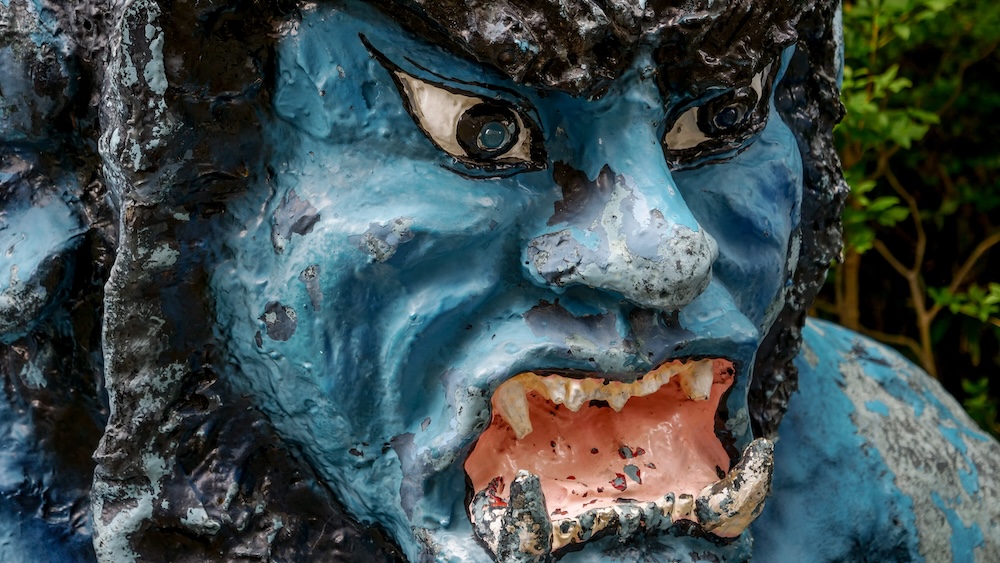 Estatua de un demonio guardián oni azul en Noboribetsu Onsen, Hokkaido, Japón: primer plano del rostro feroz y sus colmillos Primer plano de una estatua desgastada de un demonio oni azul en Noboribetsu Onsen, Hokkaido, Japón, con expresión feroz, boca rosada muy abierta, colmillos puntiagudos, ojos saltones y pelo oscuro esculpido sobre un fondo verde desenfocado.