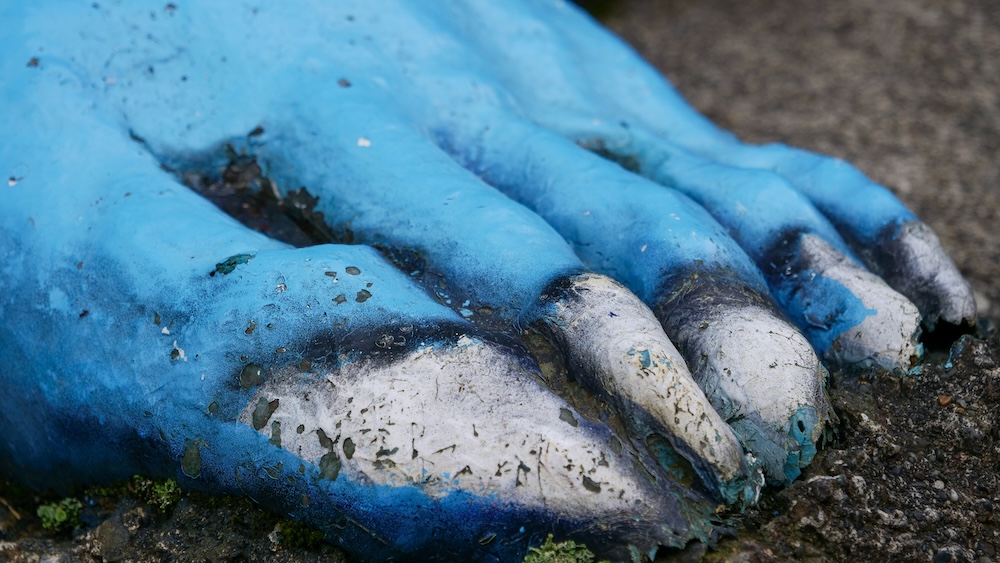 Close-up of the oversized blue foot of an oni demon statue in Noboribetsu Onsen, Hokkaido, Japan, showing thick toes, chipped white-painted claws, weathered blue paint and patches of exposed surface near the ground.