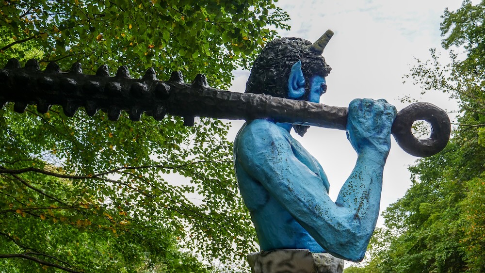 Oni azul gigante con un garrote negro de púas sobre el hombro en Noboribetsu Onsen, Hokkaido, Japón Vista lateral de una estatua gigante de oni azul en Noboribetsu Onsen, Hokkaido, Japón, que lleva un largo garrote negro con púas apoyado sobre el hombro, lo sostiene con una mano y muestra un cuerno puntiagudo entre los árboles verdes.