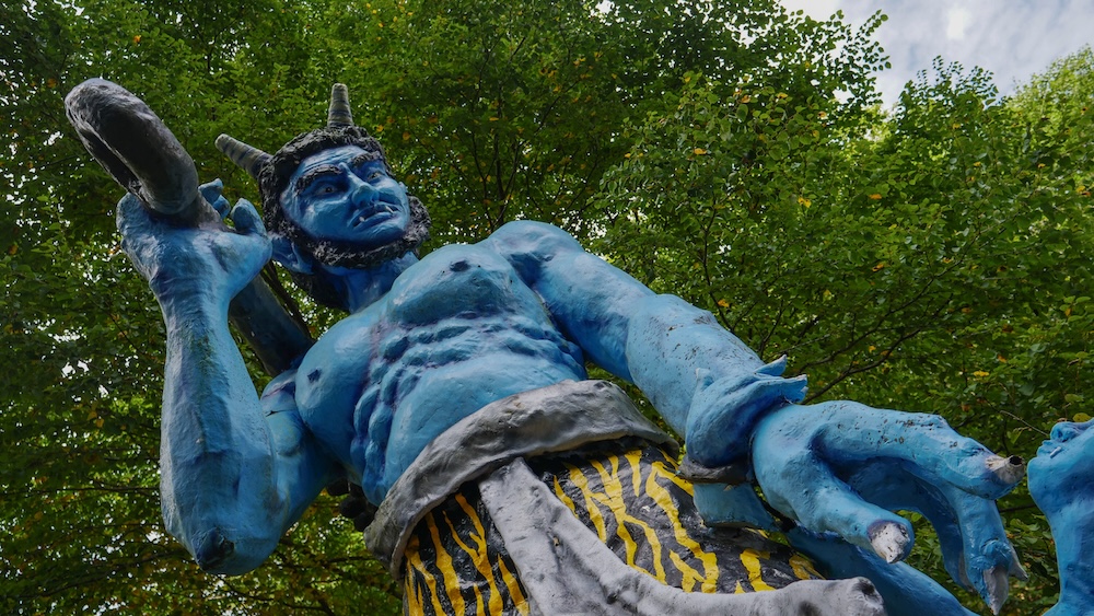 Estatua gigante de un oni azul con cuernos y garrote de púas en Noboribetsu Onsen, Hokkaido, Japón Primer plano desde abajo de una enorme estatua de un oni azul en Noboribetsu Onsen, Hokkaido, Japón. Se destacan sus cuernos, colmillos, cuerpo musculoso, garrote levantado y vestimenta amarilla con rayas de tigre frente a árboles verdes.