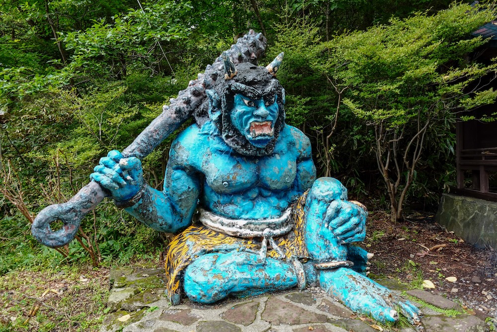 Estatua de un demonio oni azul sentado con un garrote de púas en Noboribetsu Onsen, Hokkaido, Japón Una gran estatua de un demonio oni azul sentado sobre una base de piedra en Noboribetsu Onsen, Hokkaido, Japón, con colmillos, cuernos, taparrabos amarillo con estampado de tigre y un enorme garrote con púas, rodeado de árboles verdes.