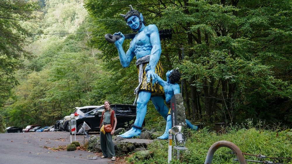 Audrey Bergner junto a un oni azul gigante y otro más pequeño al borde de una ruta en Noboribetsu Onsen, Hokkaido, Japón Audrey Bergner junto a una enorme estatua de un oni azul con cuernos y un garrote de púas, con un demonio azul más pequeño detrás, al borde de una ruta rodeada de bosque y una zona de estacionamiento en Noboribetsu Onsen, Hokkaido, Japón.