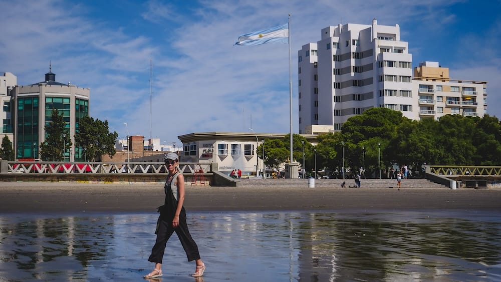 Waterfront and beach, Puerto Madryn, Chubut, Argentine Patagonia — Audrey Bergner Audrey Bergner walking along the wet shoreline in Puerto Madryn, Chubut, Argentine Patagonia, with the Argentine flag and waterfront buildings visible behind her.