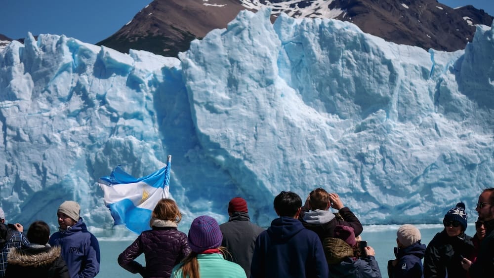 Boat passengers viewing Perito Moreno Glacier's ice wall, El Calafate, Santa Cruz, Argentina A crowd of boat passengers, one holding an Argentine flag, photographs the towering blue ice wall of Perito Moreno Glacier from the water near El Calafate, Santa Cruz Province, Patagonia, Argentina.