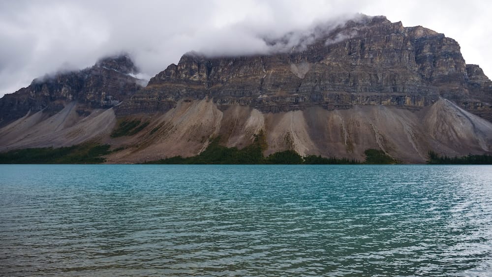 Low cloud clings to the jagged peak above Bow Lake's turquoise glacial water in Banff National Park, Alberta, along the Icefields Parkway, with dark scree slopes descending to the treeline.