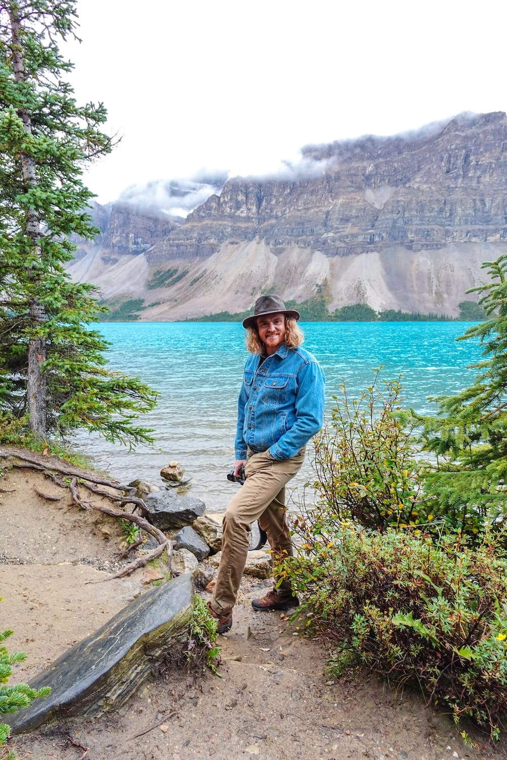 Samuel Jeffery está en la costa rocosa de Bow Lake, junto a la Icefields Parkway en Alberta, Canadá, con agua turquesa, pinos y nubes bajas sobre las montañas detrás.